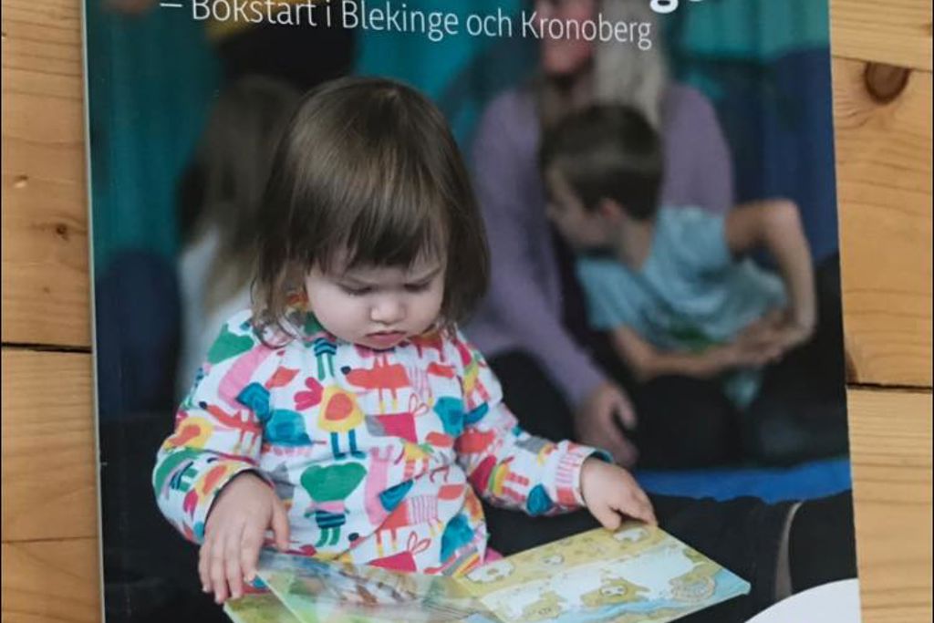 Skriften Att nå varenda unge - Bokstart i Blekinge och Kronoberg har ett läsande barn på omslaget och ligger på ett trägolv.