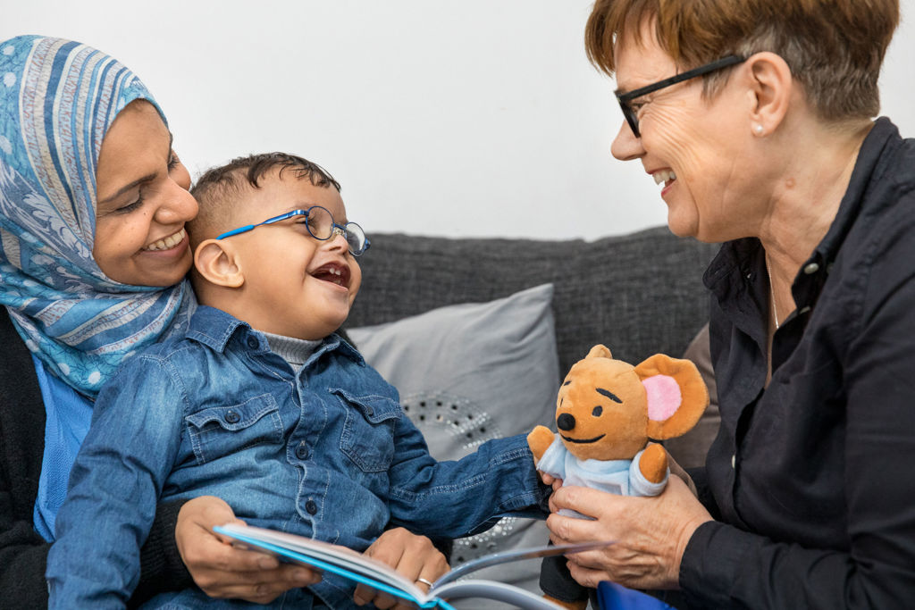 Mamma håller barn i famnen som ler och håller i en bok. Pedagog tittar glatt på dem. 